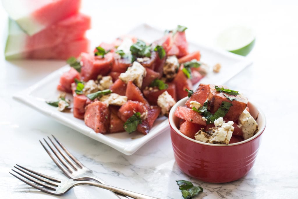 watermelon salad plated on white rectangle plate with two forks, and bowl of watermelon basil salad