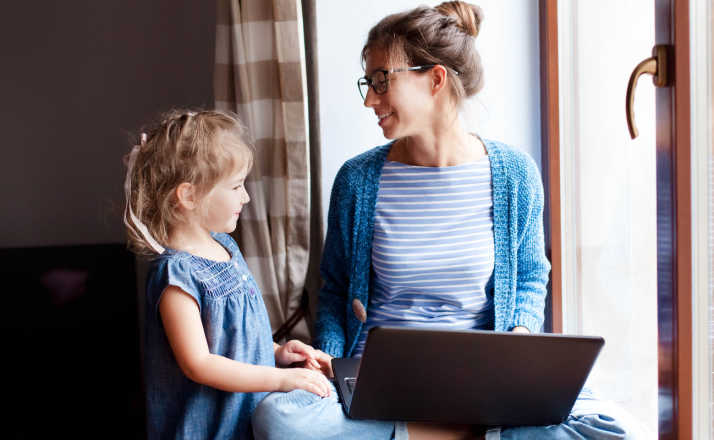 mom with glasses and laptop looking at young daughter while sitting in windowsill get a break from kids