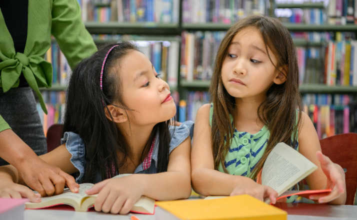 Two cute girls are jealous of each other while reading books in library while teacher teaching. People lifestyles and education. Young friendship and Kids relationship in school concept. Daycare theme