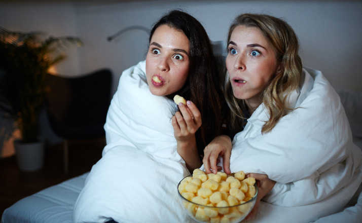 Curious girls eating corn rolls from bowl while watching scary family movies at night 