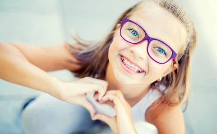 Smiling little girl in with braces and glasses showing heart with hands. When do kids get braces